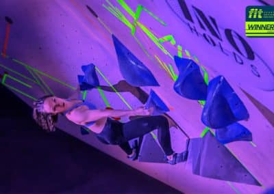 Une compétitrice grimpe en dévers sur les prises AlpX de Youkaïdi et InoHolds. Les prises sont fixées sur un mur d'escalade en salle lors d'une compétition